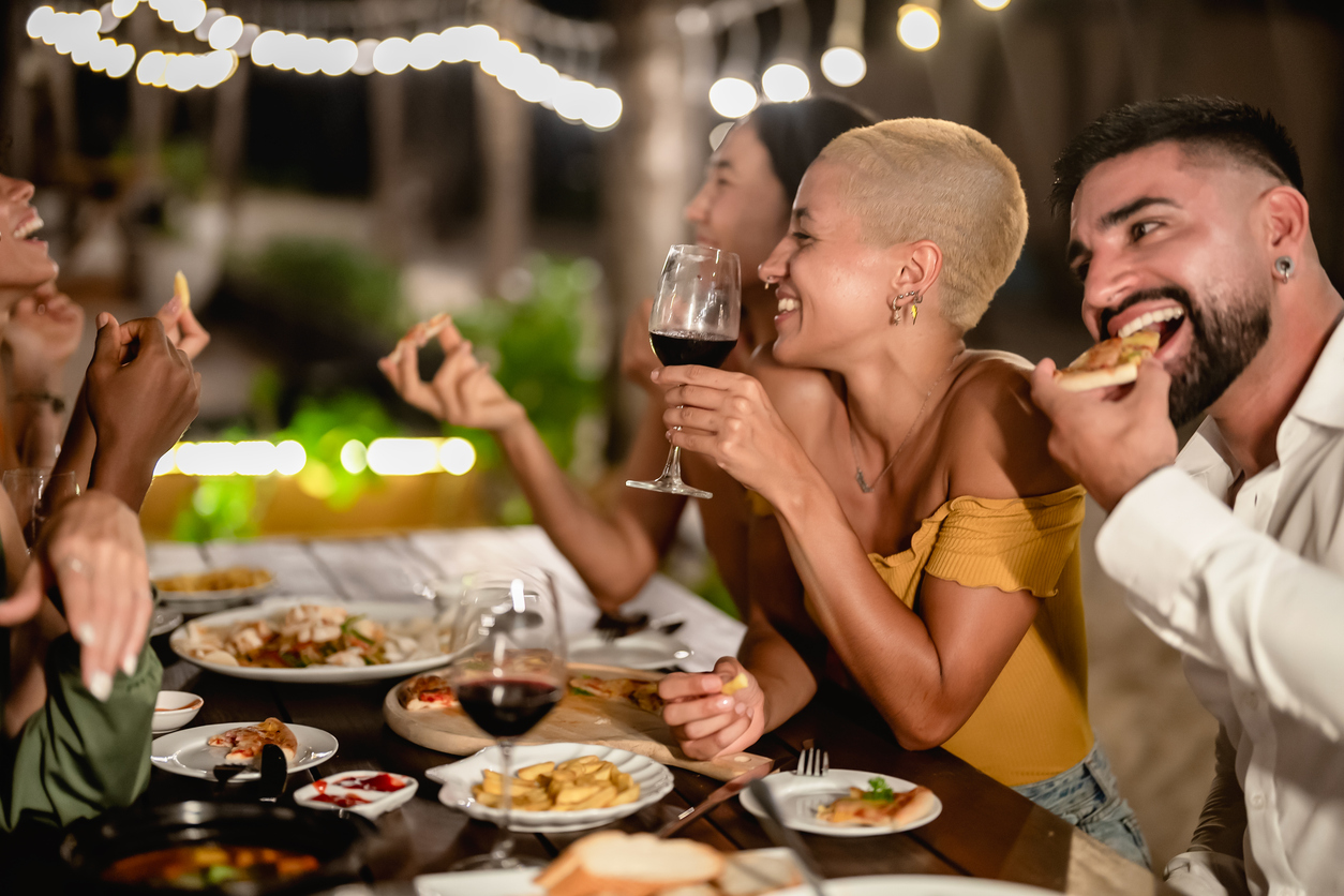 Friends laughing over wine and food at a candlelit outdoor dinner with string lights